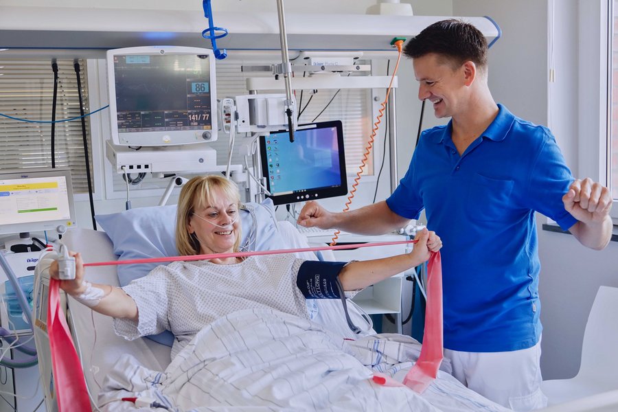 Eine Frau liegt in einem Krankenbett und macht Kraftübungen mit einem Gummiband. Daneben steht ein Pfleger und demonstriert die Ausführung der Übung.