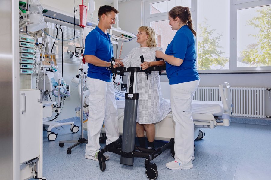 Eine blonde Frau in einem Nachthemd steht an einer Gehilfe in einem Krankenhauszimmer. Links und rechts neben ihr stehen ein Krankenpfleger und eine Krankenpflegerin und lächeln ihr aufmunternd zu.