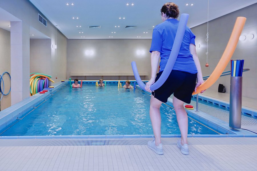 Eine Frau steht vor einem Pool und demonstriert mit zwei Poolnudeln eine Übung. Im Wasser sind drei Damen, die ihr nachmachen.