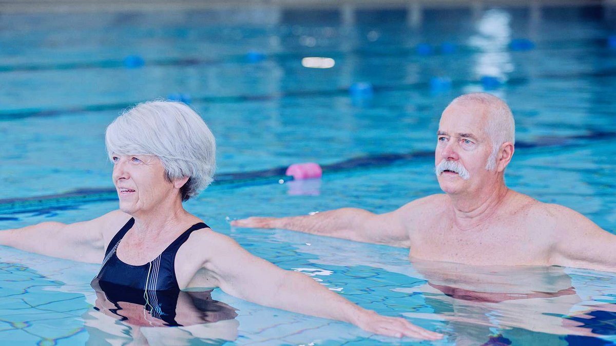 Eine ältere Dame und ein älterer Herr stehen in einem Schwimmbecken. Sie haben beide Arme nach außen ausgestreckt und blicken zu jemandem außerhalb des Sichtfeldes der Kamera.