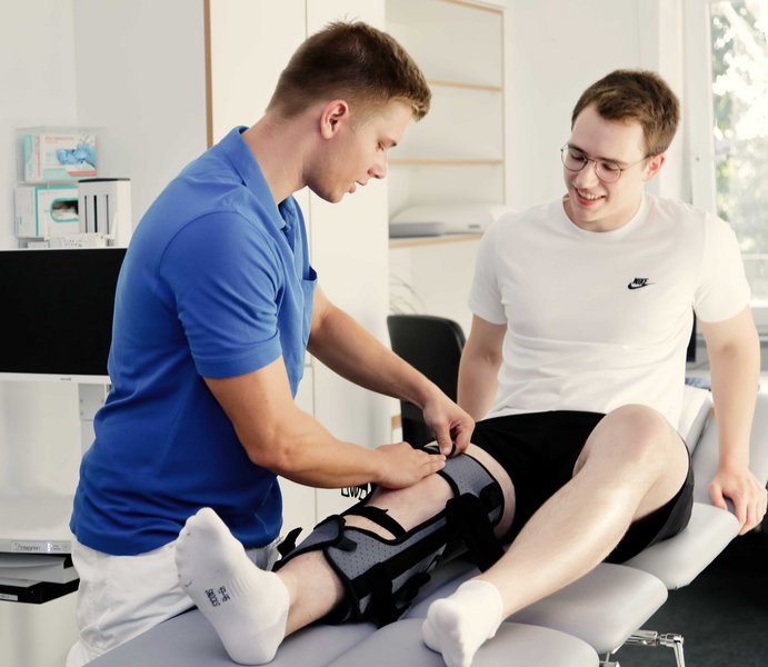 Ein Mann sitzt auf einer Liege und bekommt von einem Krankenpfleger eine Schiene um sein rechtes Bein gelegt.