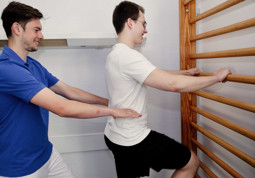 Ein junger Mann steht mit einem Bein angehoben vor einem Holzgerüst und hält sich daran fest. Hinter ihm steht ein Pfleger und assistiert ihm.