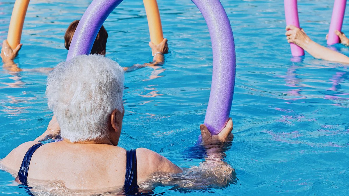 Eine ältere Dame und noch mindestens zwei weitere Personen stehen mit einer gebogenen Poolnudel in den Händen im Wasser.