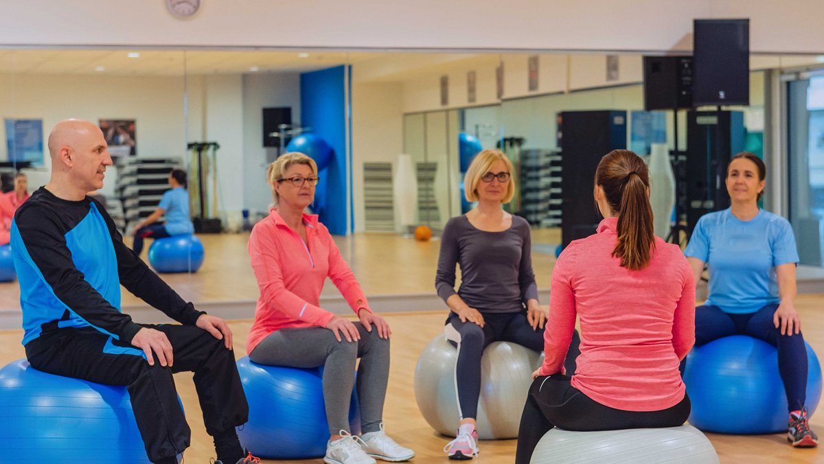 Eine kleine Gruppe sportlich gekleideter Menschen sitzt auf Medizinbällen. Eine Frau sitzt mit dem Rücken zur Kamera, die Anderen schauen Sie an.