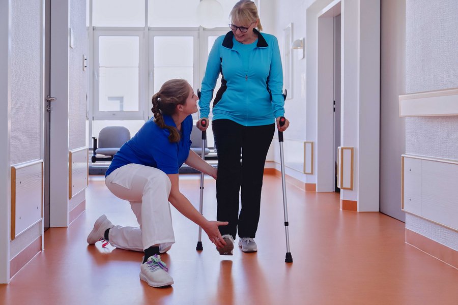 Einer Frau auf Krücken wird von einer neben ihr knieenden Pflegerin geholfen zu gehen.
